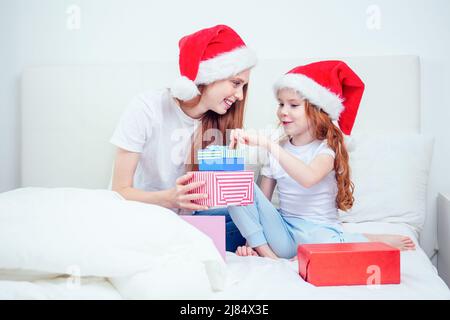 beautiful redhaired ginger woman with santa claus hat and happy little girl in pajamas sitting on bed with white pillow and blanket next to gifts Stock Photo