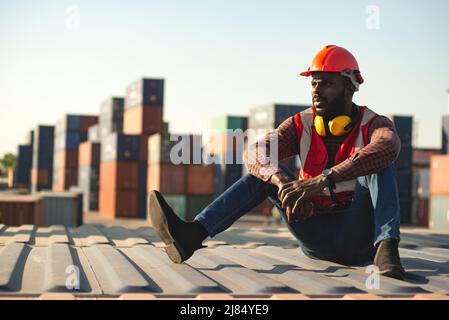 African American male technician or engineer. Sit near a container and look tired and sleepy or unemployed. Logistics industrial cargo mover concept. Stock Photo