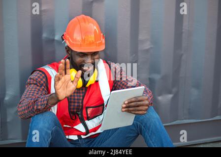 Close up happy smiling male African American engineer wearing safety vest and helmet, sitting and using mobile phone at logistic shipping cargo contai Stock Photo