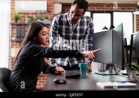 Asian application developer pointing out cloud database processing algorithm error while uploading network information. Multiethnic tech software engineers decrypting retrieved stolen data. Stock Photo