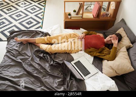 Above view of tired young woman in cardigan lying with closed eyes on bed with laptop and holding hand on forehead, work burnout concept Stock Photo