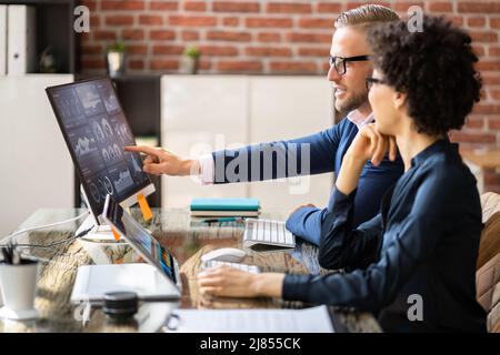 Side View Of Two Stock Market Brokers Discussing Graphs On Computer At Workplace Stock Photo