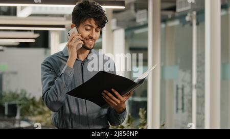 Busy arab man businessman worker in office workspace talking mobile phone answer call consulting clients discussing financial issues holds folder Stock Photo