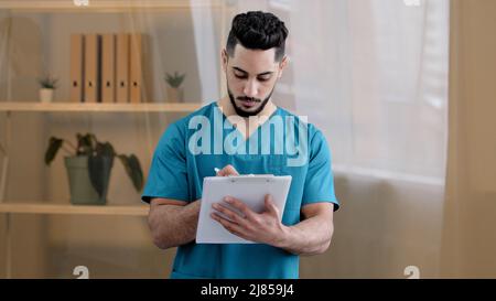 Hispanic physician male doctor man therapist medic wear uniform standing in hospital clinic writing notes fill medical form insurance notice detailed Stock Photo