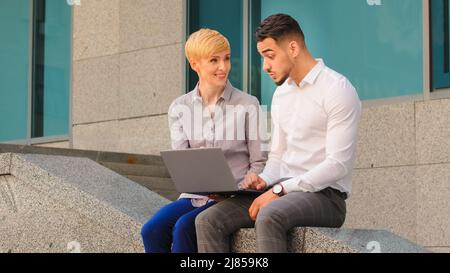 Cheerful multiethnic multiracial couple colleagues arabic hispanic man and caucasian woman standing near business center at street outdoors and Stock Photo