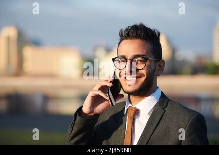 Portrait of smiling confident young Arabian businessman in eyeglasses talking by cellphone outdoors Stock Photo