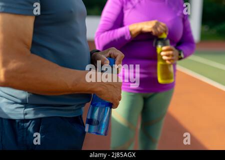 Midsection of biracial senior man and woman opening water bottles while standing in tennis court Stock Photo