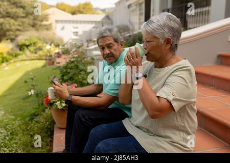 Biracial senior woman drinking coffee and senior man using digital tablet while sitting on steps Stock Photo