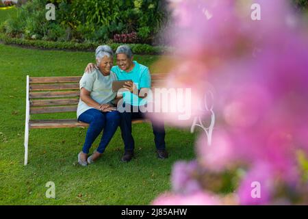Cheerful biracial senior man with wife using digital tablet while sitting on bench in park Stock Photo