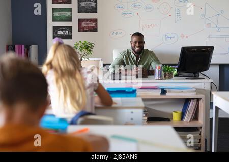 Portrait of african american young male teacher sitting at desk with students studying in classroom Stock Photo