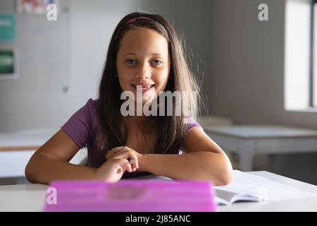 Portrait of smiling caucasian elementary schoolgirl sitting at desk in classroom Stock Photo