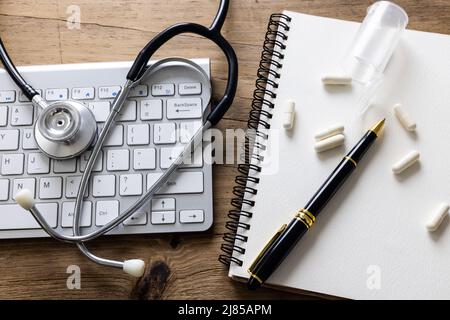 Close-up of spiral book, pen, medicines with stethoscope and computer keyboard on wooden table Stock Photo