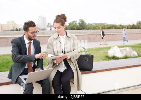 Young interracial business colleagues sitting on bench and analyzing papers using data on laptop outdoors Stock Photo