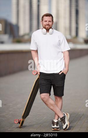 Portrait of smiling handsome bearded man holding hand in pocket and leaning on skateboard in city Stock Photo