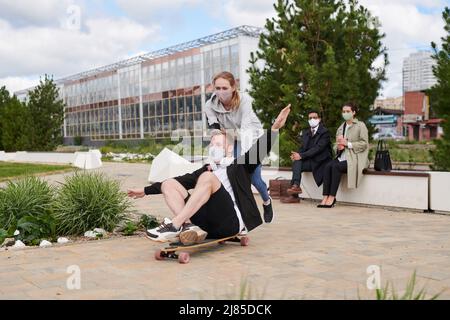 Positive young friends in cloth masks enjoying skateboarding in city park during coronavirus pandemic Stock Photo