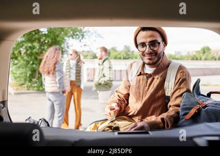 Portrait of content young Arabian guy in jacket and hat putting clothes into trunk while preparing for travel with friends Stock Photo