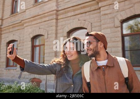 Happy young multi-ethnic couple of tourists standing against stone building and taking selfie together Stock Photo