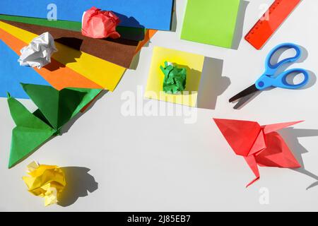 Paper crafts, scissors, colored paper sheets and scraps, on white table. Origami. Children working place. Preschooler paper artwork. Top view. Stock Photo