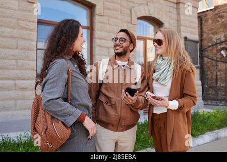 Group of positive young multi-ethnic friends checking phones and chatting with each other while spending time together outdoors Stock Photo