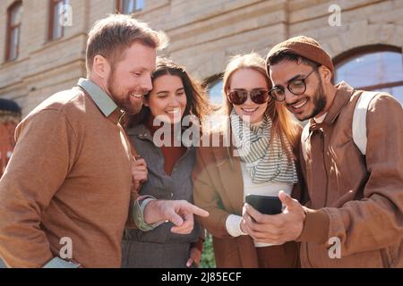 Group of cheerful young multi-ethnic friends using smartphone app outdoors while discussing cafe choice Stock Photo