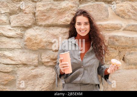 Portrait of smiling curly-haired girl in gray shirt having break for coffee and enjoying doughnut and cappuccino outdoors Stock Photo