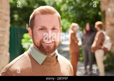 Portrait of smiling handsome young bearded college teacher in sweater standing against campus Stock Photo