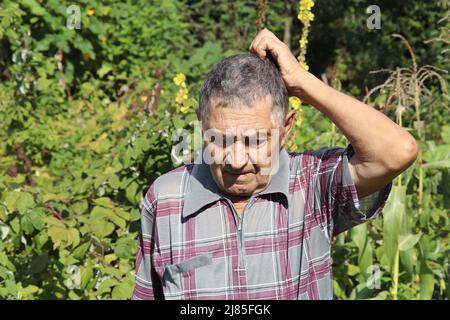 Portrait of elderly man scratches his head standing in summer garden. Concept of life in village, old age Stock Photo