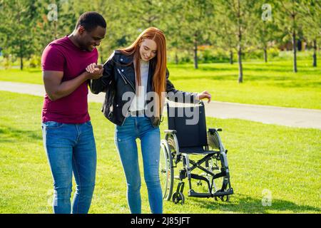 african american man helping to her redhaired ginger woman to making first steps after long recovery.she walking without her wheelchair in spring park Stock Photo