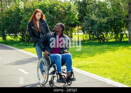 afro american man sitting on wheelchair ,his redhaired ginger girlfriend rolling stroller in autumn park. having romantic date Stock Photo