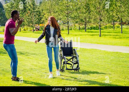 african american man helping to her redhaired ginger woman to making first steps after long recovery.she walking without her wheelchair in spring park Stock Photo