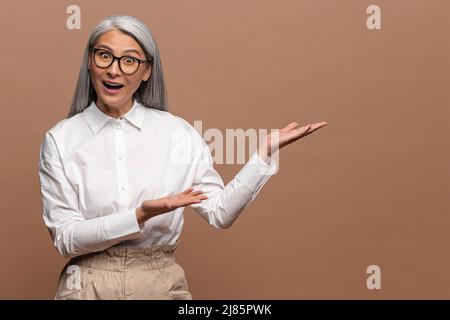 Portrait of surprised mature woman pointing finger and palm right at empty space, looking at camera with open mouth and shocked expression. Indoor studio shot isolated on beige background Stock Photo