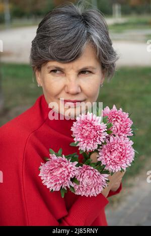 Semi-profile portrait of attractive gray-haired woman in a red blouse with a bouquet of purple chrysanthemums,looking to a side. Waist up shot. Stock Photo