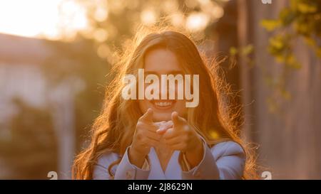 Portrait of female model stands on street in sunlight sunset happy smiling woman girl looking at camera makes gesture hey you hands pistols points Stock Photo