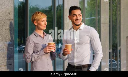 Two multi-ethnic business partners multiracial colleagues arabic spanish man and caucasian woman standing outdoors near company building drinking Stock Photo