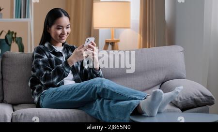 Asian girl woman sitting on sofa at home relaxing watching news feed on social media choosing clothes online shop internet store dating site wi-fi Stock Photo