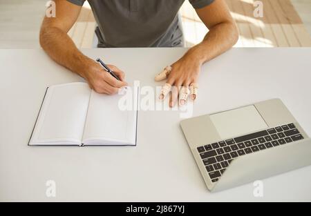 Man wearing braces on his broken fingers sitting at desk and writing in notebook Stock Photo