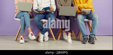 Young people with laptops and notebooks sitting in row on chairs on background of purple wall. Stock Photo