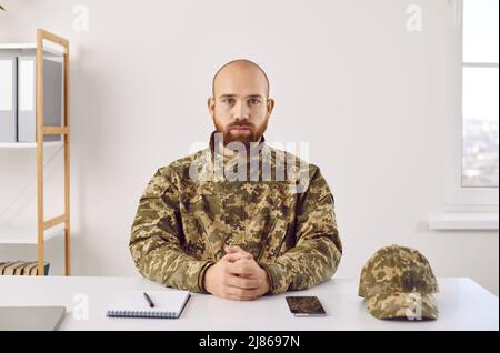 Portrait of serious soldier in camouflage uniform sitting at office desk and looking at camera Stock Photo