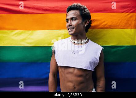 Smiling young gay man in stylish clothes standing against pride flag Stock Photo