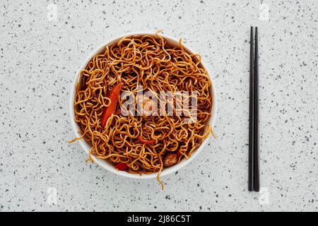 high angle view of an off-white plate with some chicken yakisoba noodles and a pair of black chopsticks placed on a white mottled stone surface Stock Photo