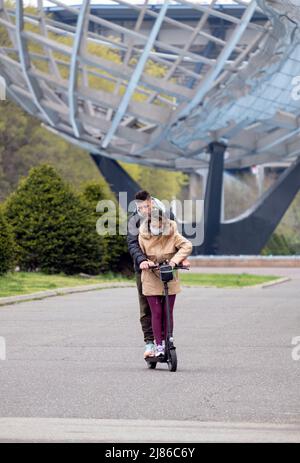 On a chilly spring morning, an attractive couple ride an electric scooter at Flushing Meadows Park with the Unisphere in the background. Stock Photo