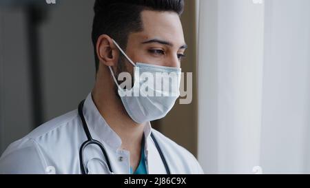 Male pensive thoughtful arab man therapist surgeon wear white uniform medical face mask standing at workplace looking out window preventing spread Stock Photo