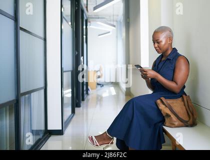 A young Black woman texts while sitting and waiting in office corridor Stock Photo
