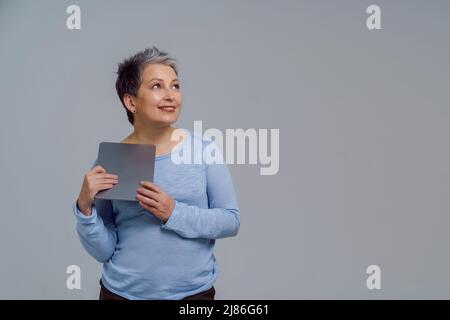 Dreaming beautiful mature grey haired woman in 50s holding digital tablet working or shopping online or checking on social media. Pretty woman in blue blouse isolated on white background. Toned image. Stock Photo