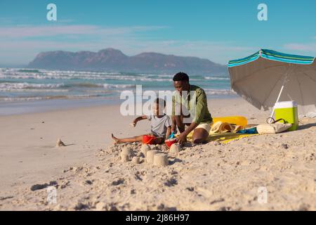 African american young man making sand castles with son at beach against sea and sky on sunny day Stock Photo