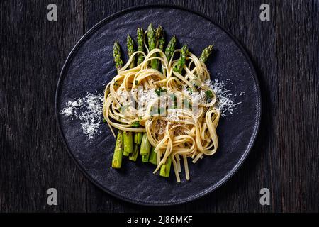 roasted asparagus spears with spaghetti sprinkled with shredded pecorino cheese and parsley on black plate on dark wooden table, horizontal view from Stock Photo