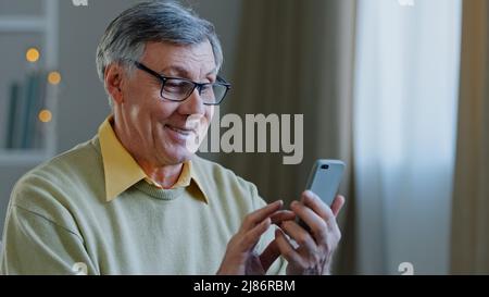 Mature elderly mature 60s caucasian man with phone male grandfather uses mobile app at home buys medicine online brows on net writes message to Stock Photo