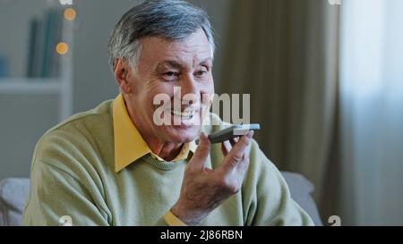 Mature grey-haired 60s man 70s senior old male at home hold smartphone speak loud to phone microphone speaker audio voice message using speakerphone Stock Photo