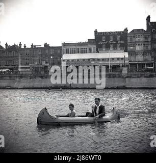 1950s, historical, a father and son in a canoe on the calm sea at Margate, Kent, England, UK. In the distance, on the high street seafront, a tearoom of the famous Briitsh company, J. Lyons & Co, the leading tea and restuarant chain of the era. Founded in 1884, J Lyons was famous for its 'corner house' tea rooms and ther uniformed waitresses, who were known as 'nippies'. On one of the buildings a sign for the company and another promoting their 'Swiss Rolls'. Stock Photo