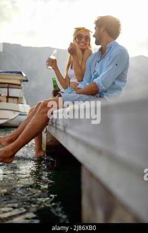 Boyfriend and girlfriend with drink, sitting, laughing and having small talk on wooden jetty by sea and boat pier. Tourism, togetherness, lifestyle co Stock Photo